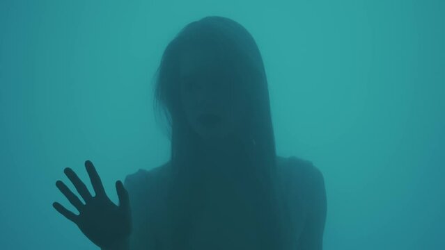 Portrait of female in neon light behind the glass window in steam and water drops. Girl puts hands on glass, looks sideways, worried face.
