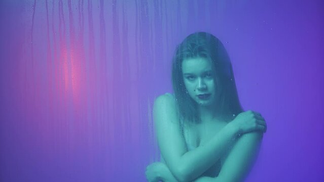Portrait of female in neon light behind the glass window in steam and water drops. Girl with makeup and natural hairstyle touching smooth skin.
