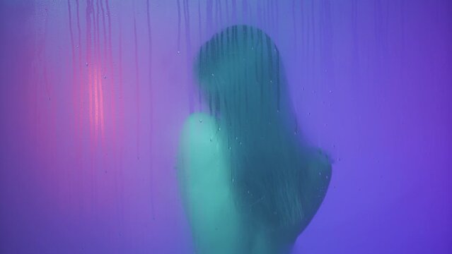 Portrait of female in neon light behind the glass window in steam and water drops. Girl with long hair posing with back at camera, touching skin.