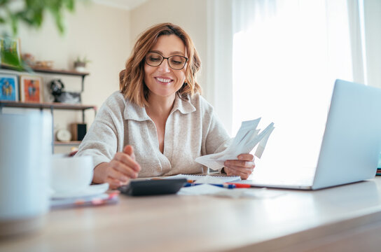 Beautiful middle-aged woman in glasses and paper bills joyfully laughing while calculating revenue grows at home office. Small business, successful investment, bull market, money savings concept image