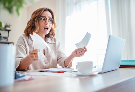 Beautiful middle-aged woman in glasses extremely surprised higher family expenses gazing at paper bills and laptop home office. Small business troubles, utilities costs or money savings concept image