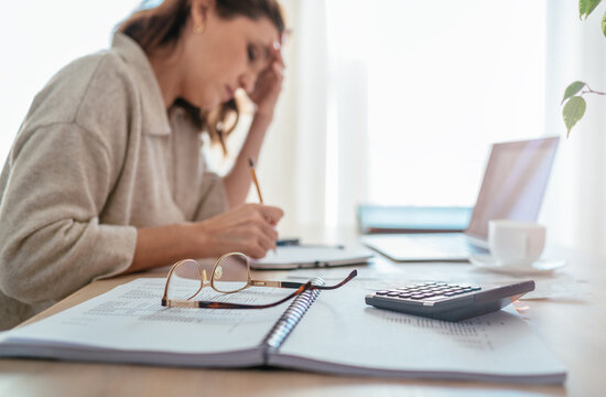 Eyeglasses and calculator close-up and tired accountant woman making marks in paper documents with modern laptop sitting background next bright daylight window. Business, home finances money savings