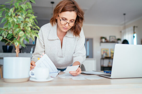 Beautiful middle-aged woman in glasses thinking about family business budget gazing at monthly bills and documents at home living room office. Small business home finances, money savings concept image