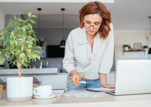 Beautiful middle-aged woman in glasses thinking about family business budget gazing at monthly bills and documents at home living room office. Small business home finances, money savings concept image