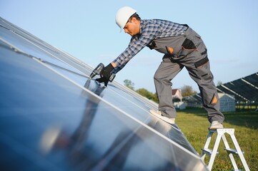 Portrait of Young indian man technician wearing white hard hat standing near solar panels against...