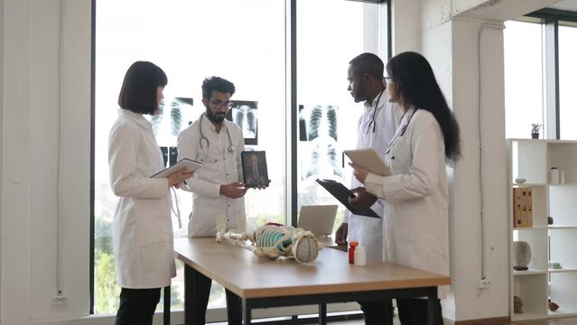 Young adult male teacher in class, holding x-ray of spine on tablet and teaching human skeleton anatomy, using artificial skeleton model to explain structure of bones and joints for group of students.