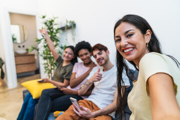 Multiracial friends sharing a joyful moment while taking a group selfie on the couch, creating memories at home.