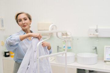 Cheerful middle-aged female doctor walks into office, takes robe off coat hanger and puts it, smiling looking camera. Professional practitioner getting ready to work in clinic. Concept of healthcare.