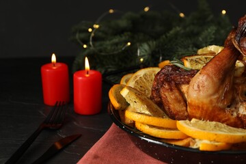 Baked chicken with orange slices and burning candles on black table, closeup