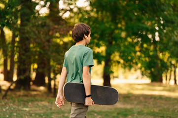 Back view shot of a teen boy walking while holding his skateboard outdoors in a park.