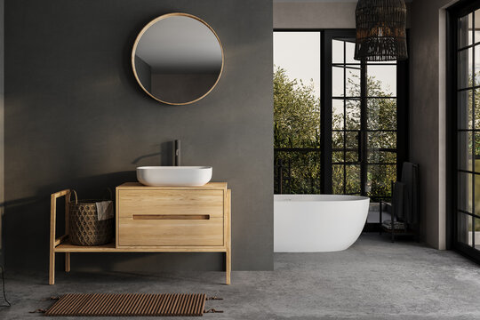 Modern bathroom interior with dark walls, ceramic basin with mirror, bathtub and grey concrete floor. Minimalist black bathroom with modern furniture and big windows, yard.