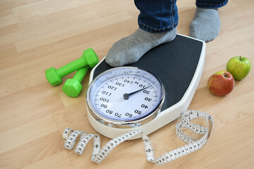 Feet of a man in gray socks stepping on an analog weight scale, green dumbbells, measuring tape and apples lying nearby on a wooden floor, fitness and diet concept, copy space