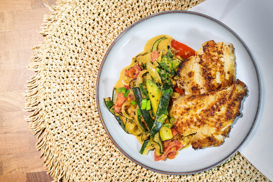 Two fried redfish fillets on vegetables such as zucchini, tomatoes and leek, on a gray plate and a woven natural fiber placemat, healthy recipe for low carb diet, top view from above, copy space