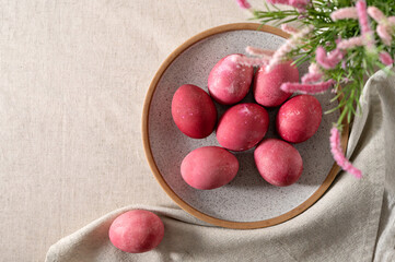Pink colored eggs on plate, on neutral beige linen tablecloth background with blurred spring flowers and soft natural sun light, aesthetic Easter holiday backdrop, copy space