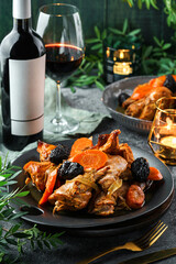 Holiday baked chicken in honey with dried prunes and carrots in plate served with glasses, bottle of wine, candles, plants, festive decoration. Christmas and New Year food, bokeh, light