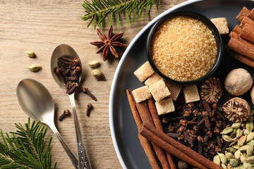 Plate with different aromatic spices, spoons and fir branches on wooden table, flat lay