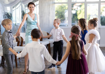 Cheerful junior schoolchildren in party dresses having fun dancing in circle with female pedagogue - obrazy, fototapety, plakaty