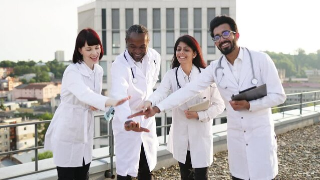 Group of multi ethnic medical specialists doctors looking at camera while stacking their hands together for successful cooperation, standing outdoors on rooftop of hospital during break.