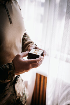 A military soldier in uniform holds a box with gold wedding rings in his hands while standing by the window. Hands close up. Copy space.