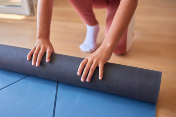 High-angle view of sporty preteen girl rolling up yoga mat after training at home by window in...