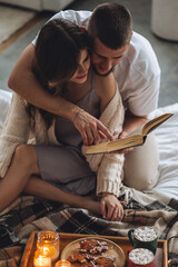 Happy young couple relaxing together in the bedroom at home, reading book. Man and woman enjoying lazy cozy weekend at home, embracing, kissing, cuddling. Simple pleasures, domestic life