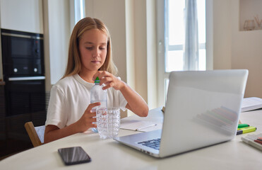 Thirsty little schoolgirl writing in exercise book, taking break and drinking fresh cool filtered...