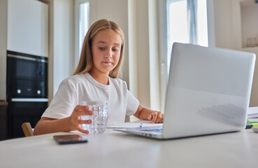 Thirsty little schoolgirl writing in exercise book, taking break and drinking fresh cool filtered...