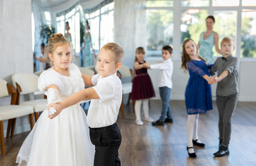 Joyful schoolboys and schoolgirls of primary classes training valse dance in events salon