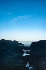 Tidal pool reflecting the blue sky, merging into the blue ocean water.  With the Moon high in the...