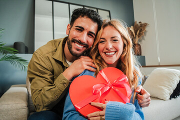 Portrait of a real young caucasian loving couple smiling and looking at camera with a valentine heart shaped gift. Handsome husband embracing her wife holding a romantic velentines day present. High