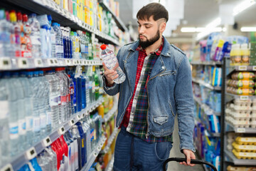 Portrait of focused young glad cheerful positive man purchasing bottled water in grocery store - obrazy, fototapety, plakaty