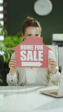 Time to move on. smiling young woman realtor in modern green office in white blouse with home for sale sign.