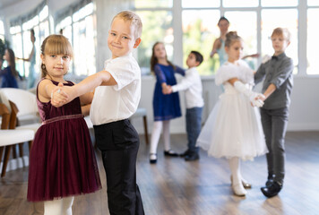 Enthusiastic preteen children, boys and girls in party attire performing elegant waltz in pairs in sunny hall of dance school with smiling female teacher in background..