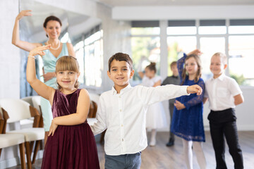 Happy interested preteen pupils preparing for festive school event, rehearsing ballroom dance steps in spacious classroom while female teacher observing from background..