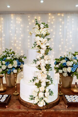A four-tiered white wedding cake decorated with white roses on a wooden table and LED lights in the background