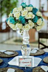 a round table decorated with a blue tablecloth, plate holder and a flower arrangement with a glass vase