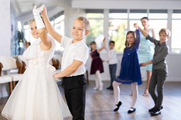Fototapeta na wymiar Enthusiastic preteen children, boys and girls in party attire performing elegant waltz in pairs in sunny hall of dance school with smiling female teacher in background..