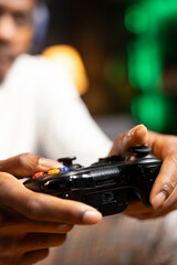 Obraz na płótnie Canvas Focus shot on controller held by gamer in blurry background playing videogames. Close up shot of gamepad used by man enjoying gaming session in living room while sitting down in front of TV