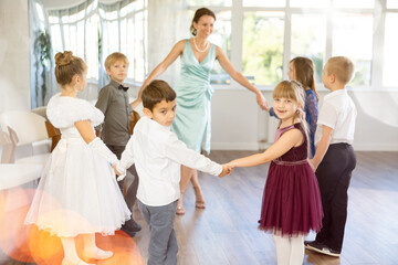 Happy little children in elegant dresses doing round dance with their teacher in school hall