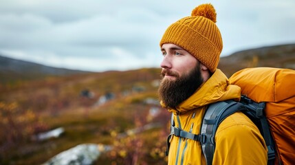 Amidst the crisp autumn air, a rugged man donning a bright yellow hat and backpack stands tall against the majestic mountain backdrop, his bearded face showcasing a sense of determination and adventu