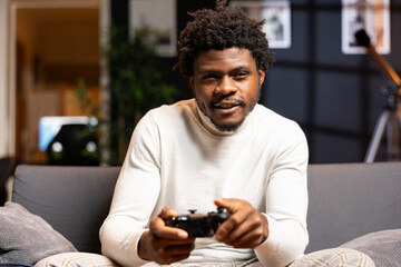 Happy man playing shooter videogame in warm apartment, holding controller. Gamer participating in online multiplayer game using gaming system, spamming buttons on gamepad - obrazy, fototapety, plakaty