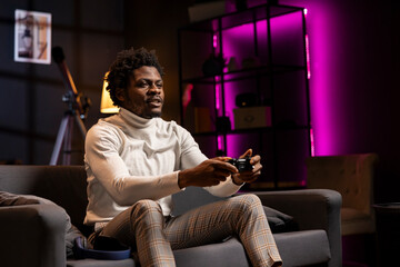 Cheerful man playing videogame in pink neon lit home theatre, holding controller. Gamer smiling,...