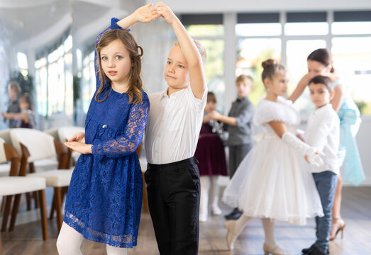 Happy little children in elegant dresses practicing waltz dance with teacher in school hall