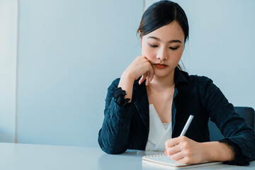 Beautiful young Asian woman writes letter on notebook while sitting at office desk. Content writer...