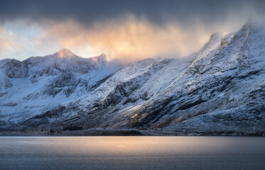 Snowy mountains in low clouds, sea bay, reflection in water at sunset in winter. Lofoten islands,...