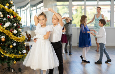 Joyful minor schoolboys and schoolgirls training valse dance in events salon adorned with Xmas tree