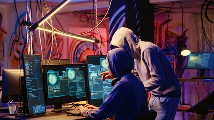Hackers in neon lit room writing lines of code on computer, having burst of joy after successfully...