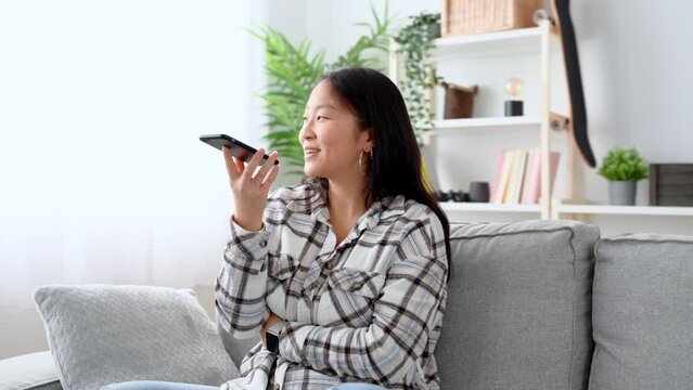 Asian young woman recording voice message on phone while sitting on sofa in the living room