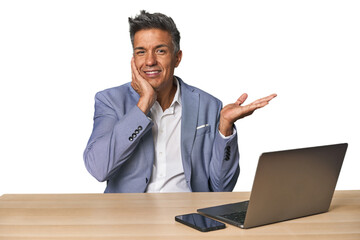 Elegant businessman at desk with laptop holds copy space on a palm, keep hand over cheek. Amazed and delighted.