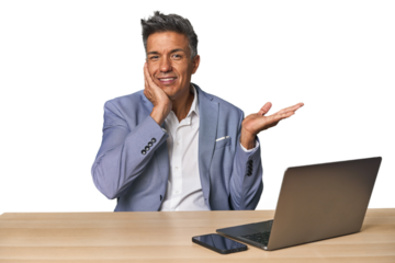 Elegant businessman at desk with laptop holds copy space on a palm, keep hand over cheek. Amazed and delighted.